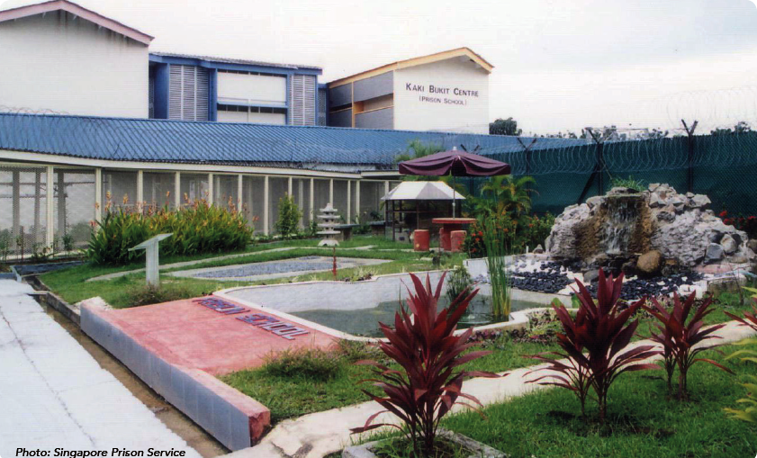 A garden with red plants, a pond, a small waterfall, and a building in the background labeled "Kaki Bukit Centre (Prison School)."
